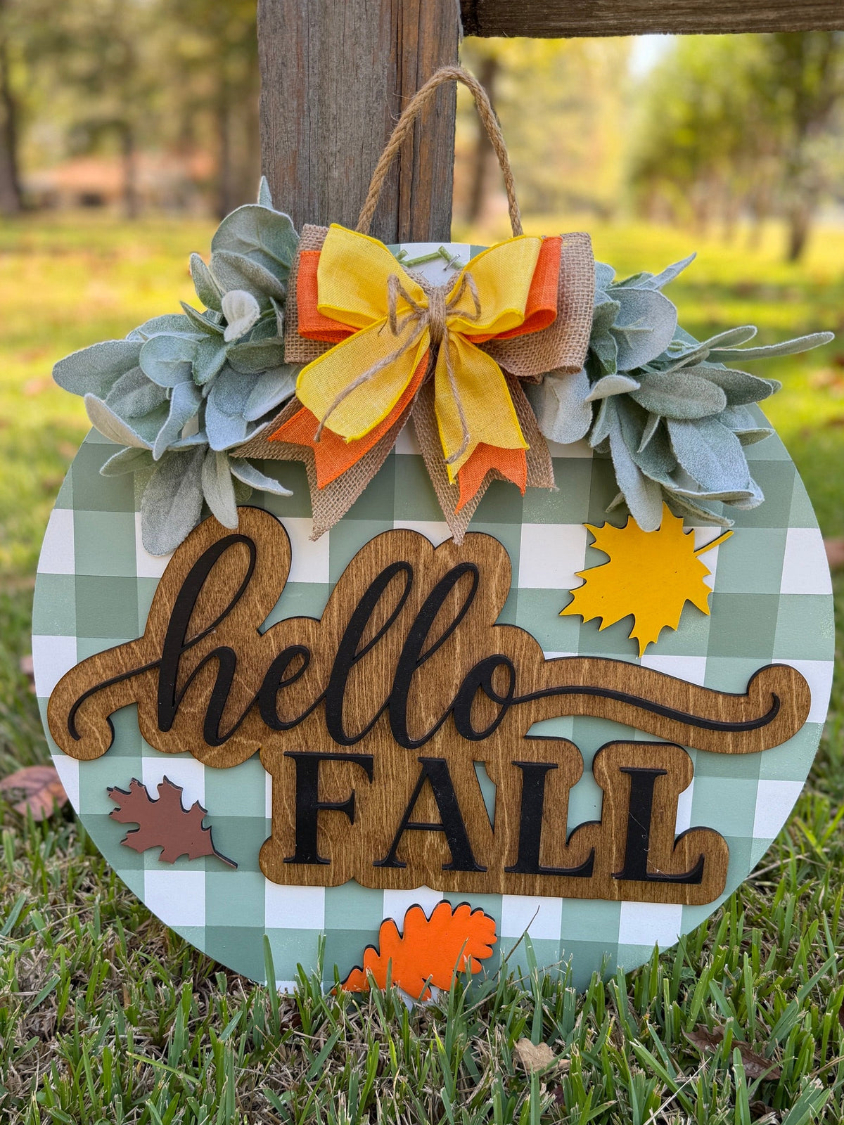 Round wooden 'hello FALL' sign with a green and white gingham pattern, decorated with faux eucalyptus leaves, a yellow and orange burlap bow, and autumn leaf accents, hanging on a wooden post in a grassy yard.